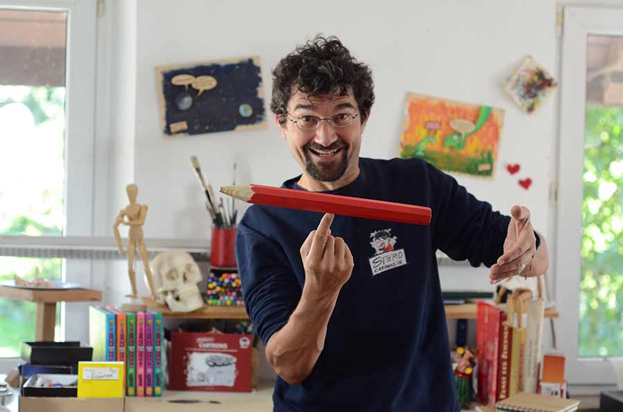 Cartoonzeichner STERO in seinem Atelier balanciert lachend einen Riesenbleistift auf dem Zeigefiger